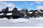 Snow-covered exterior view of a large building complex with multiple peaked roofs and a tall evergreen tree in front, under a partly cloudy blue sky.