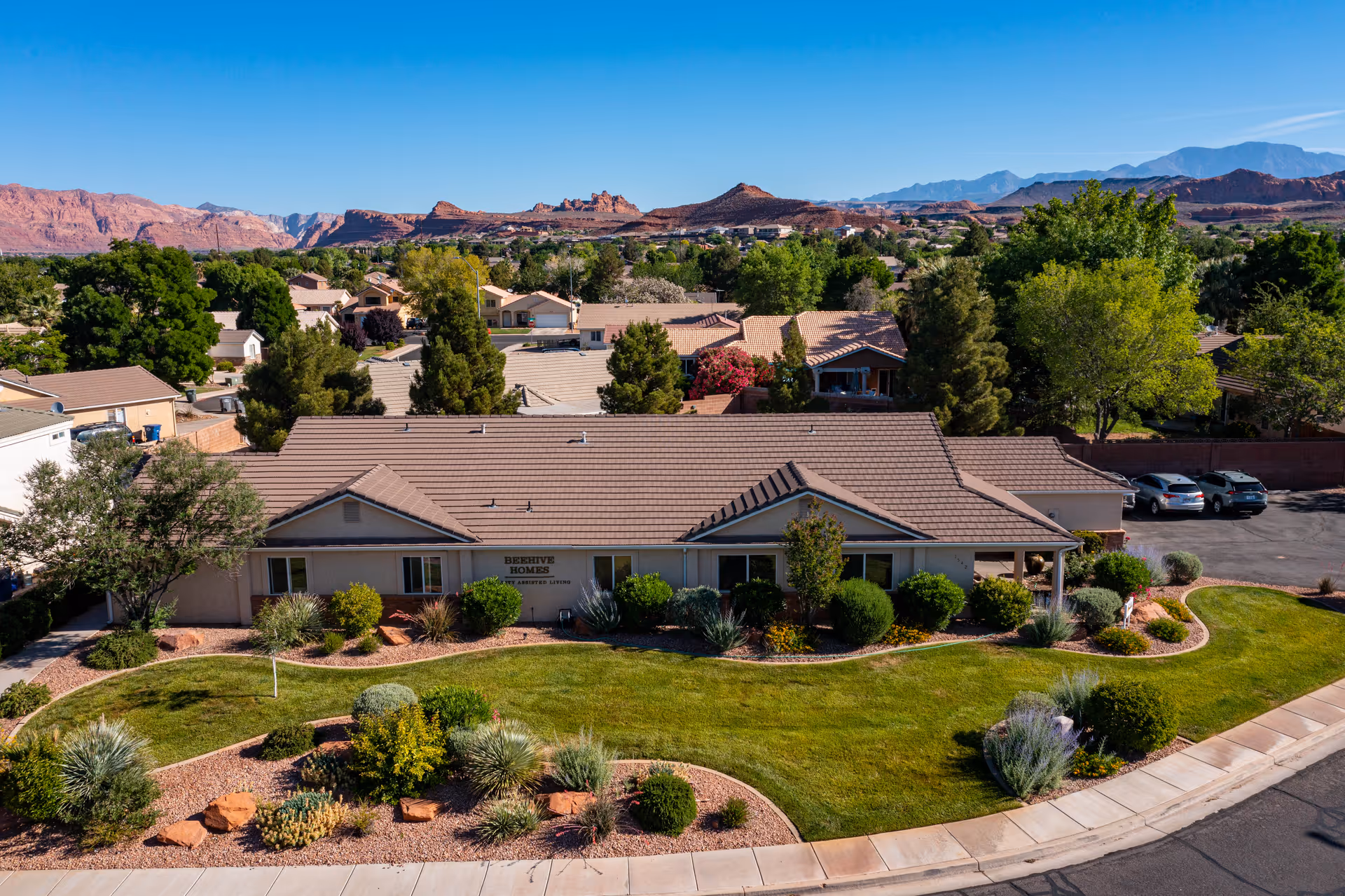 Aerial view of Beehive Homes of St George - Snow Canyon, a single-story senior living facility surrounded by well-maintained landscaping with green grass, bushes, and desert plants. The building has a tiled roof and is situated in a residential neighborhood with mountains visible in the background under a clear blue sky.