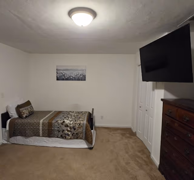 A simple bedroom with a single bed covered in a patterned brown and beige quilt, a pillow, and a cushion. There is a wooden dresser with drawers on the right side and a flat-screen TV mounted on the wall above it. A white closet door is visible next to the dresser. The walls are plain white with a framed picture of flowers hanging above the bed. The floor is carpeted in beige.