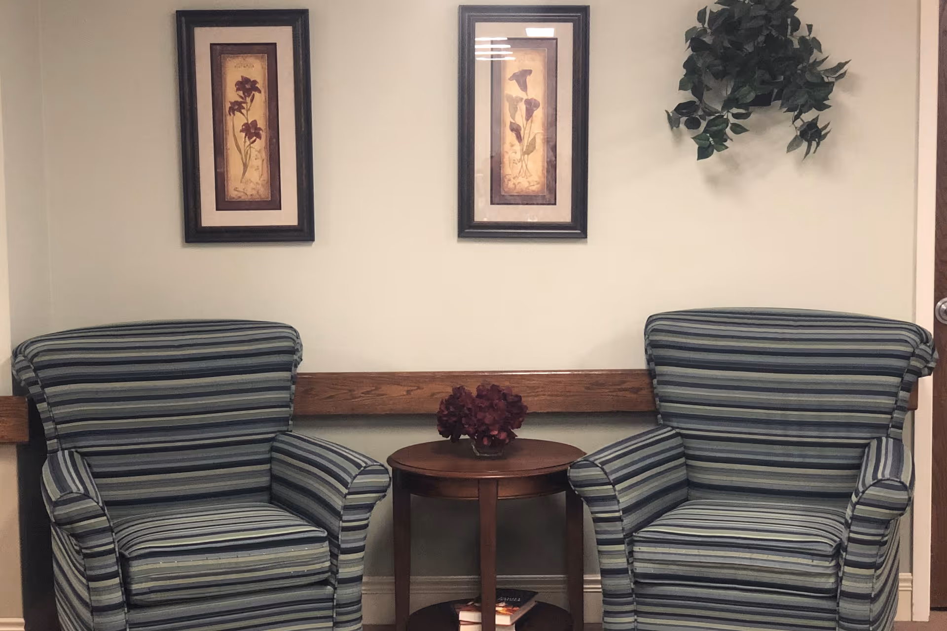 Two striped armchairs with blue and green tones are positioned on either side of a small round wooden table with a dark red flower arrangement on top. Above the chairs, two framed floral prints hang on a light-colored wall, with a small green leafy plant decoration to the right.