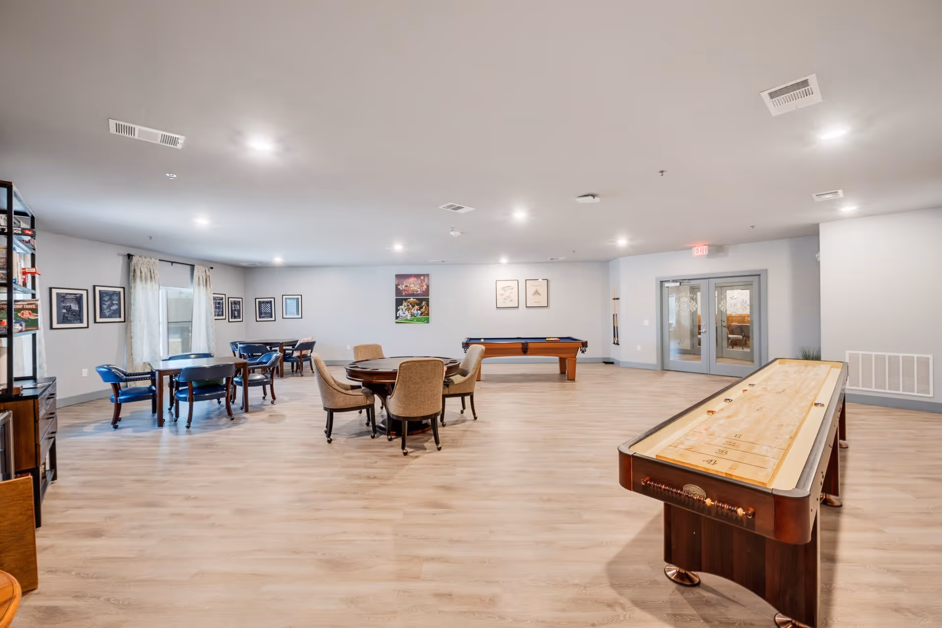 A spacious recreational room in a senior living facility featuring a shuffleboard table in the foreground, a pool table against the far wall, and several round tables with chairs arranged for games or socializing. The room has light wood flooring, light gray walls adorned with framed pictures, and recessed ceiling lights. Double glass doors are visible on the right side.