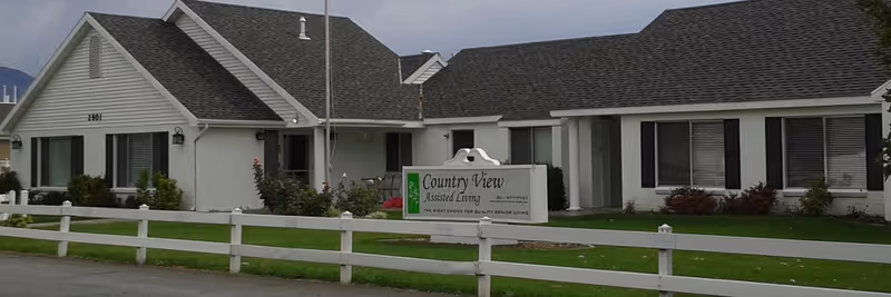 Exterior view of a single-story white building with a dark shingled roof, surrounded by a white fence and green lawn. A sign in front reads 'Country View Assisted Living' with additional text about quality service living. The sky is overcast.
