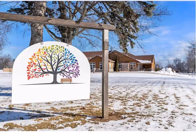 A colorful tree logo on a white hanging sign in front of a single-story brick building with snow on the ground and trees in the background under a clear blue sky.