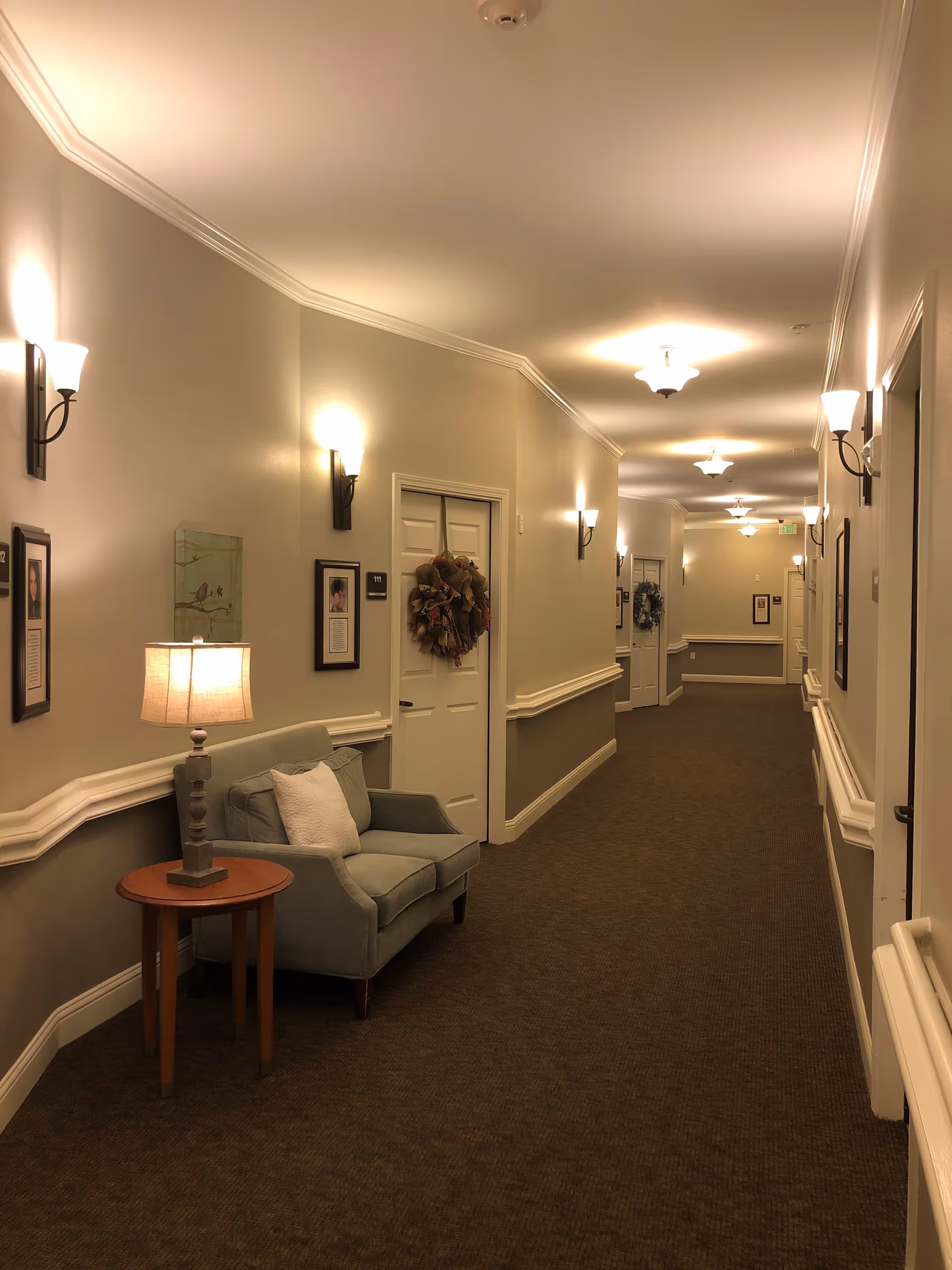 A well-lit hallway in a senior living facility with beige walls and brown carpet. The hallway features multiple white doors, some decorated with wreaths, and framed pictures on the walls. There is a small seating area with a gray armchair and a wooden side table with a lamp. Wall sconces and ceiling lights illuminate the corridor.