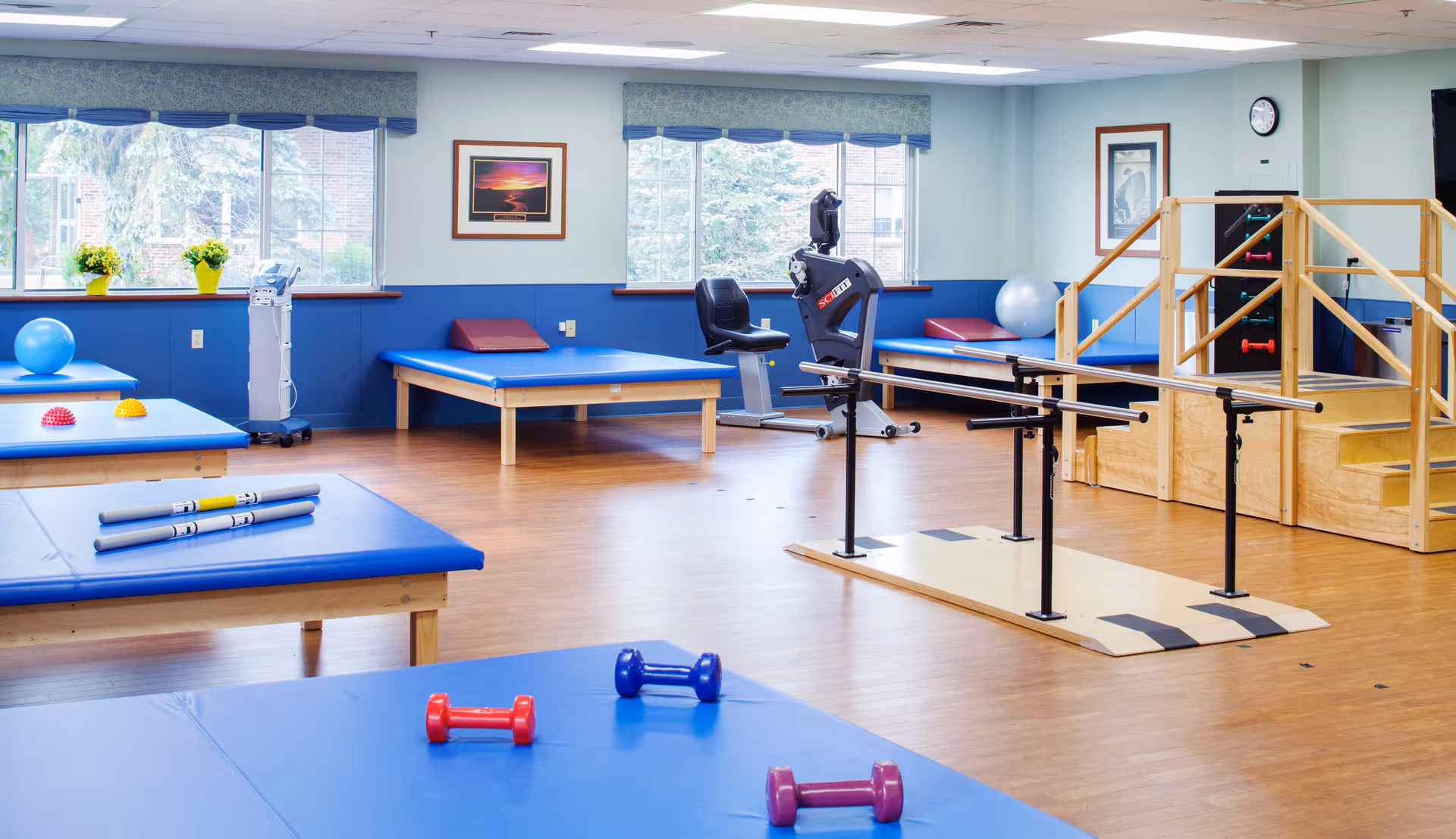 A bright physical therapy or rehabilitation room with blue padded tables, small dumbbells, exercise balls, parallel bars for walking practice, a stationary exercise bike, and a wooden staircase with handrails. Large windows let in natural light and framed pictures hang on the walls.