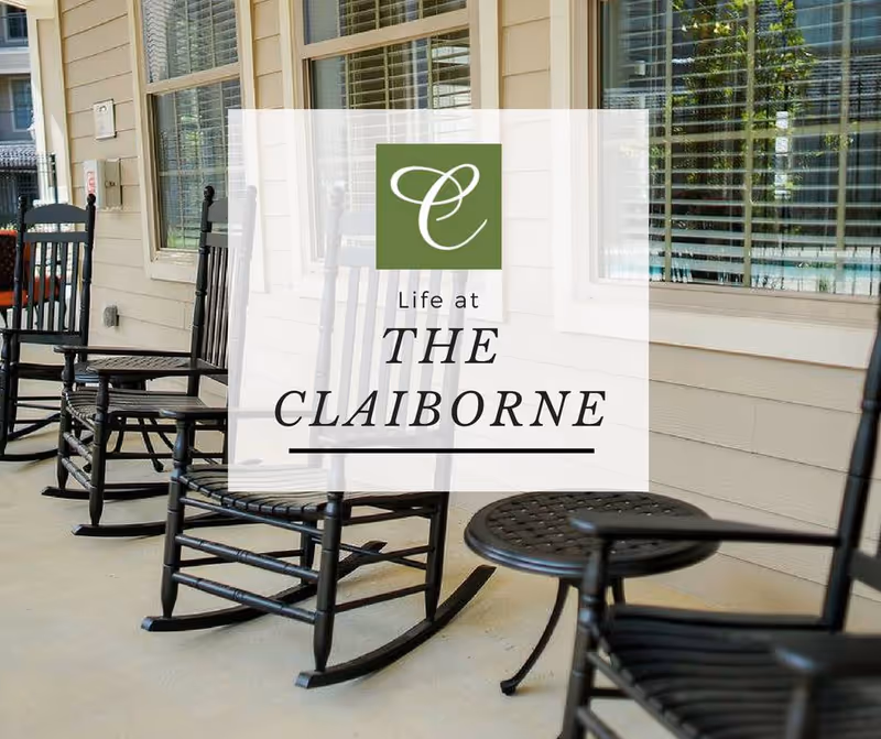 Covered front porch with black rocking chairs and small tables in front of windows and a translucent 'Life at THE CLAIBORNE' logo overlay.