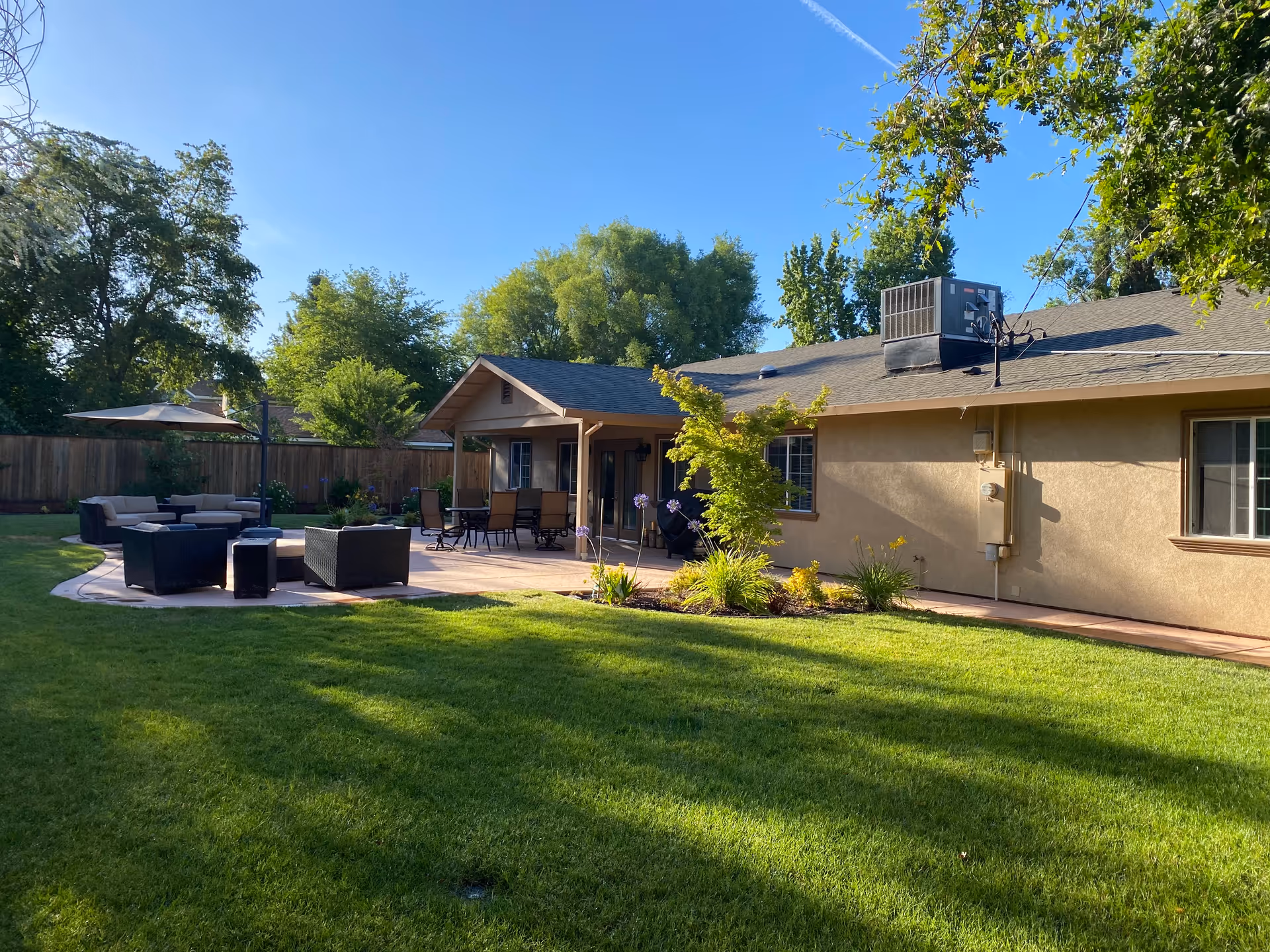 A sunny backyard with a well-maintained green lawn, patio area with outdoor seating including sofas and chairs, a table with an umbrella, and a beige single-story building with windows and an air conditioning unit on the roof. Trees and a wooden fence surround the yard.