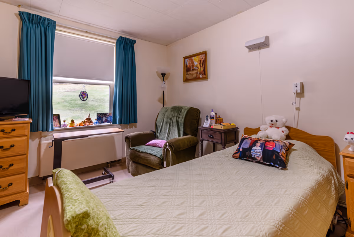A cozy bedroom in a rehabilitation and nursing center featuring a single bed with a light green quilt, a teddy bear, and a pillow with a family photo. Next to the bed is a wooden nightstand with a phone and some personal items. There is a comfortable green armchair with a purple pillow and a green throw blanket. A window with teal curtains lets in natural light, and a wooden dresser with a TV on top is positioned nearby. The walls are decorated with a framed painting and a wall-mounted light fixture.