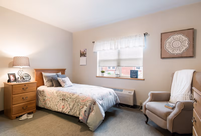 A cozy bedroom with a single bed covered in a patterned comforter and multiple pillows. Next to the bed is a wooden nightstand with a lamp, a clock, a framed photo, and a decorative 'home' sign. A pair of slippers is on the floor beside the nightstand. There is a window with blinds and a white valance, with small decorative items on the windowsill including a sign that says 'Enjoy Today.' An upholstered armchair with a white throw blanket and a book sits near the window. A framed decorative piece hangs on the wall above the chair.