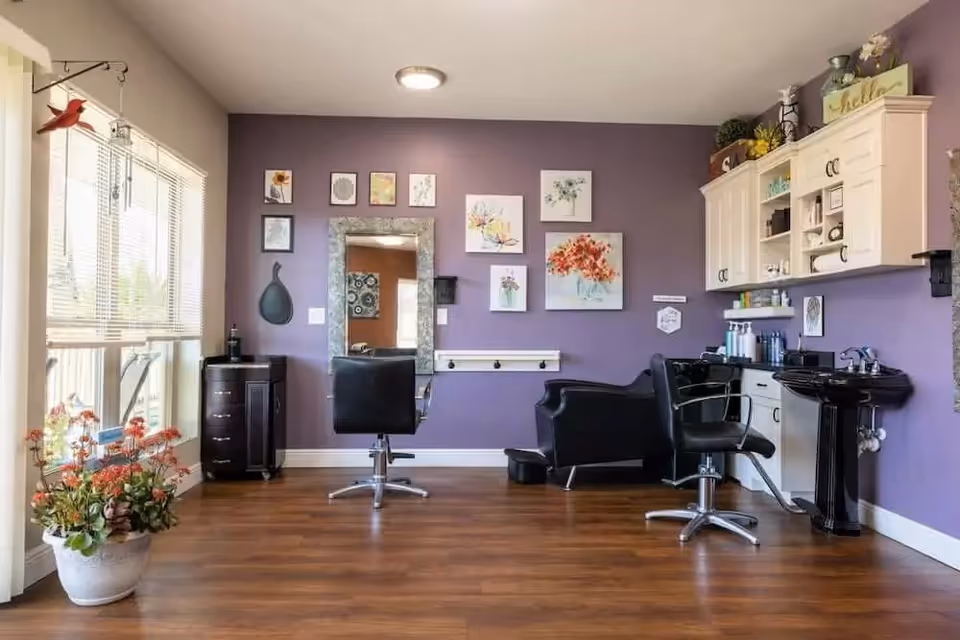 Interior of a salon area with two black salon chairs, a large mirror on a purple wall decorated with floral artwork, a black wash basin, white cabinets with shelves holding hair products, and a large window with blinds letting in natural light. There is a potted plant with orange flowers on the wooden floor near the window.