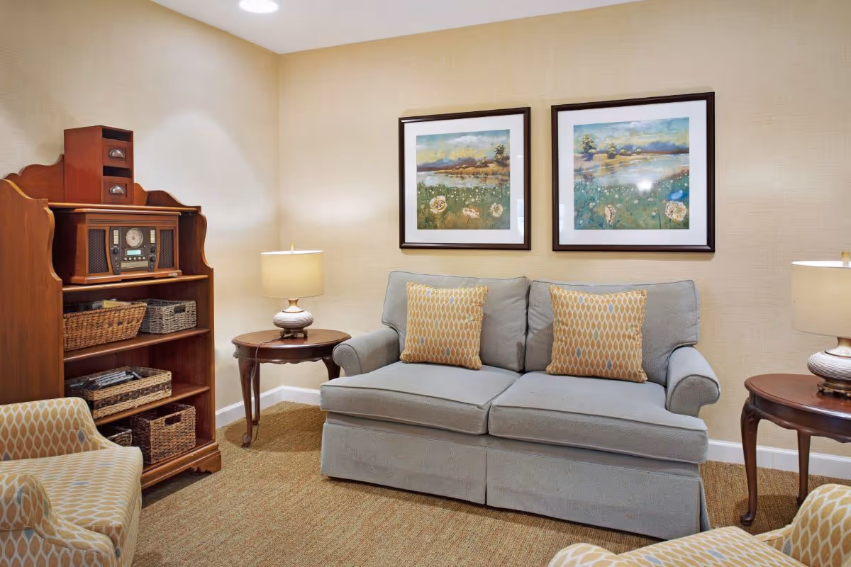 A cozy living room with a light blue sofa adorned with two patterned yellow pillows, two matching armchairs, a wooden side table with a lamp, and a wooden shelving unit holding baskets and a vintage-style radio. Two framed landscape paintings hang on the beige wall behind the sofa.