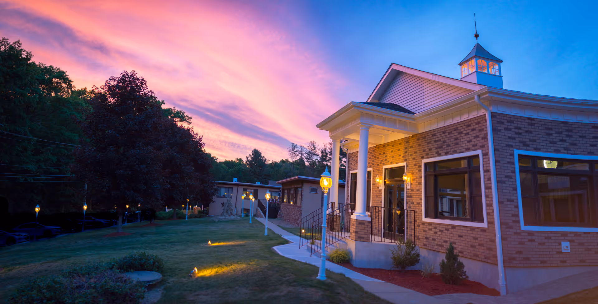 Exterior view of a brick building with white columns and large windows at dusk, with a colorful pink and purple sky in the background. The building is surrounded by a well-maintained lawn, trees, and lit lamp posts along a walkway.