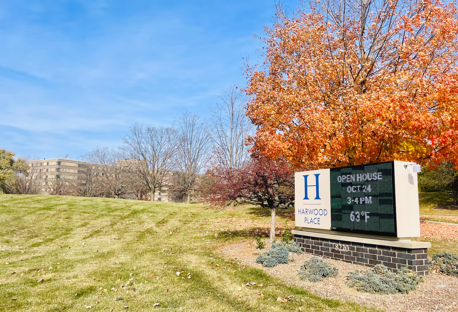 Outdoor scene showing a grassy area with a digital sign for Harwood Place Senior Living. The sign displays information about an open house event and the current temperature. Behind the sign, there are trees with autumn-colored leaves and a large building in the background under a clear blue sky.