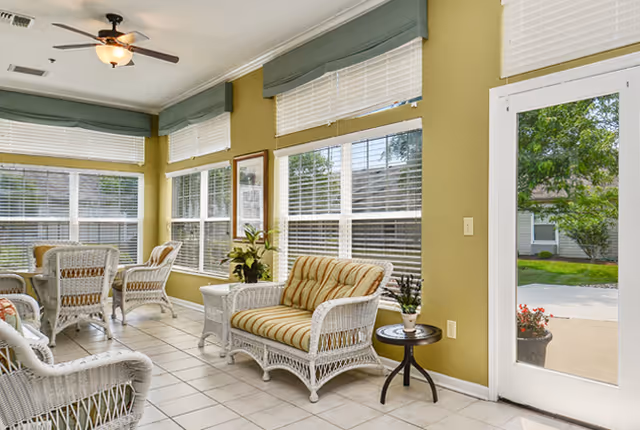 Bright sunroom with large windows and white blinds, furnished with white wicker chairs and a loveseat with striped cushions, a small round side table with a plant, and a ceiling fan with light. The room has light green walls and tiled floor, and a glass door leading outside to a patio area with greenery.