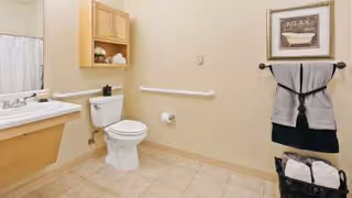 A clean and spacious bathroom with beige walls and tiled floor. The room features a white toilet, a wall-mounted sink with a large mirror above it, a wooden cabinet with towels, a towel rack holding two folded towels, a basket with rolled towels, and a framed picture on the wall.