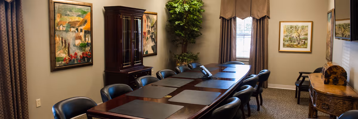 A conference room with a long wooden table surrounded by black leather chairs. The table has black desk pads and a telephone in the center. The room has beige walls adorned with framed paintings, a large window with curtains, a tall green plant in the corner, and wooden furniture including a cabinet and a side table.