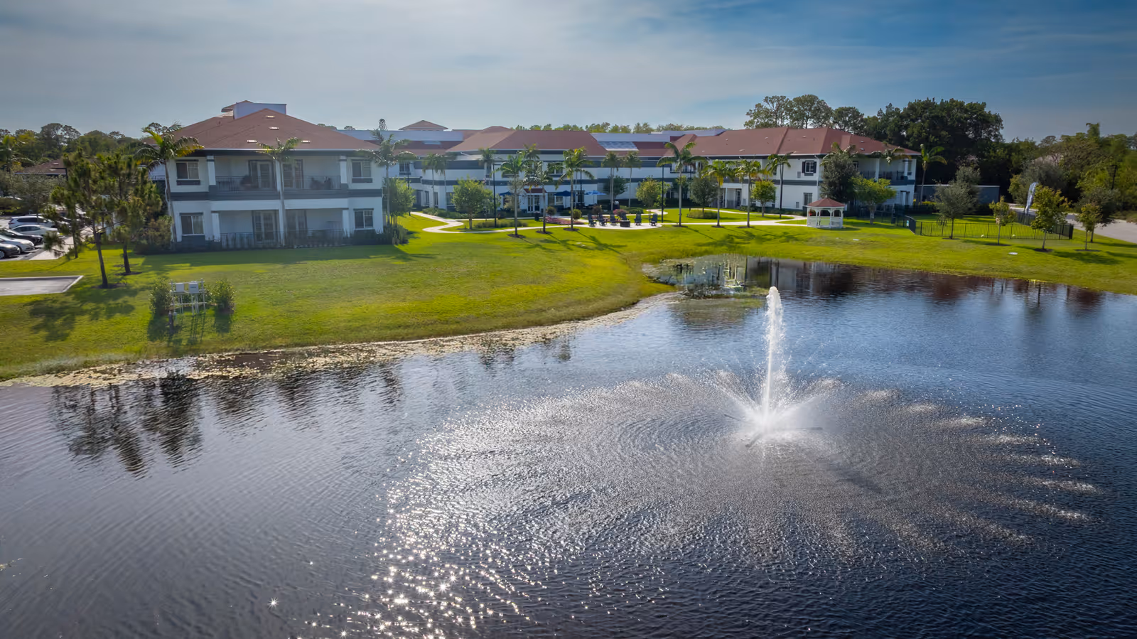 Two-story senior living buildings overlooking a pond with a central fountain and grassy grounds.