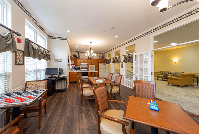A bright and spacious common area in a senior living facility featuring a kitchen with wooden cabinets, a dining area with multiple tables and chairs, and a game table with checkers. Large windows with valances allow natural light to fill the room. Adjacent to the kitchen and dining area is a lounge with comfortable seating and soft lighting.