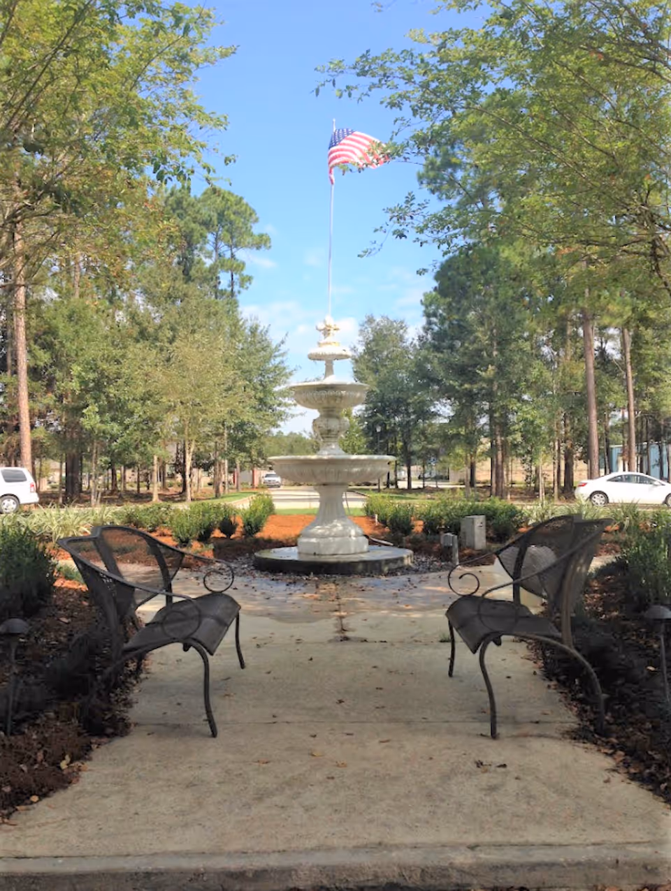 Outdoor garden area with two black metal benches facing a three-tiered white fountain topped with an American flag, surrounded by trees and parked cars in the background under a clear blue sky.