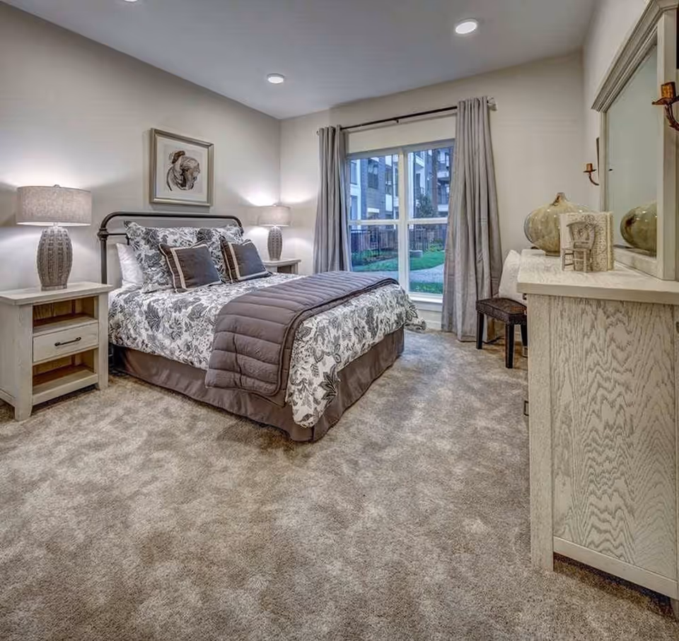 A neatly made bed with floral patterned bedding and two dark accent pillows in a bedroom. The room has beige carpet, two bedside tables with matching lamps, a large window with gray curtains showing an outdoor view of a building and greenery, and a dresser with decorative items and a mirror.