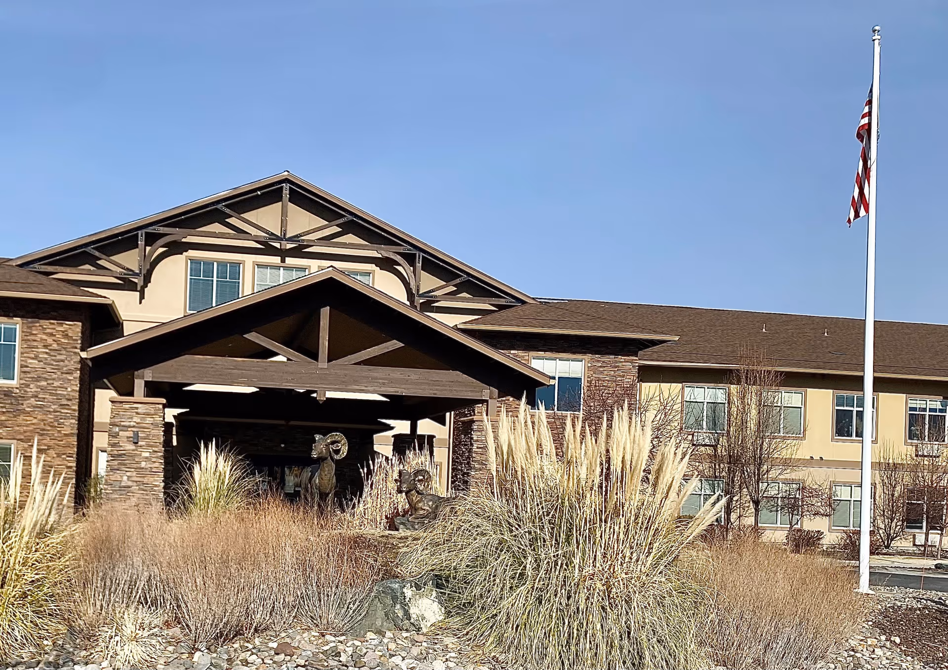 Front entrance of a senior living building with a covered porte-cochere, ornamental grasses and an American flag flying on a pole.