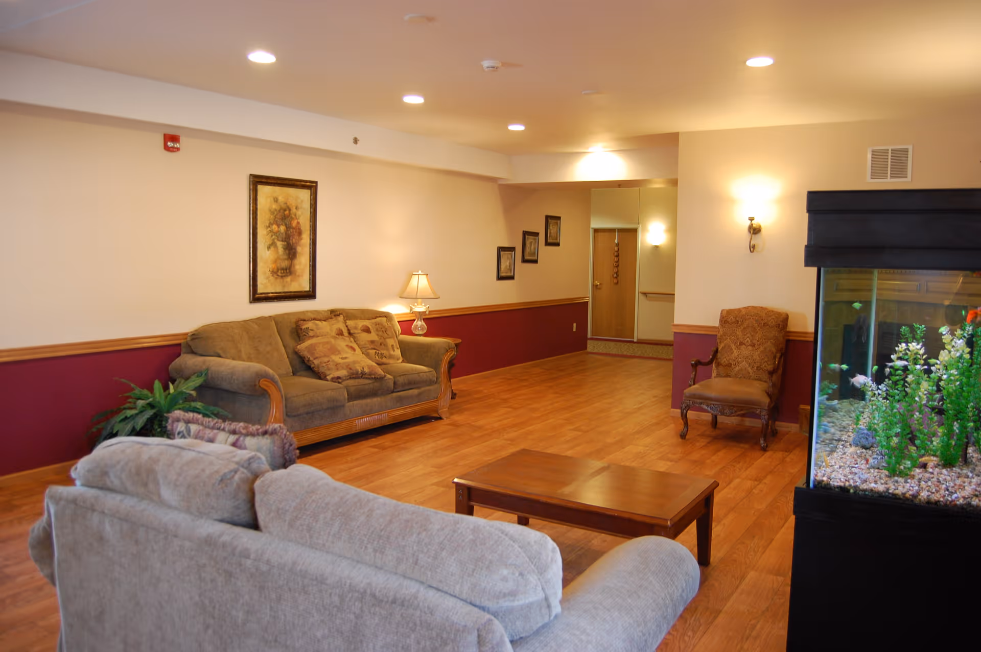 A cozy living room area in an assisted living facility featuring two sofas with cushions, a wooden coffee table, an armchair, a large fish tank with plants and fish, framed artwork on the walls, a table lamp, and warm lighting with wooden flooring.
