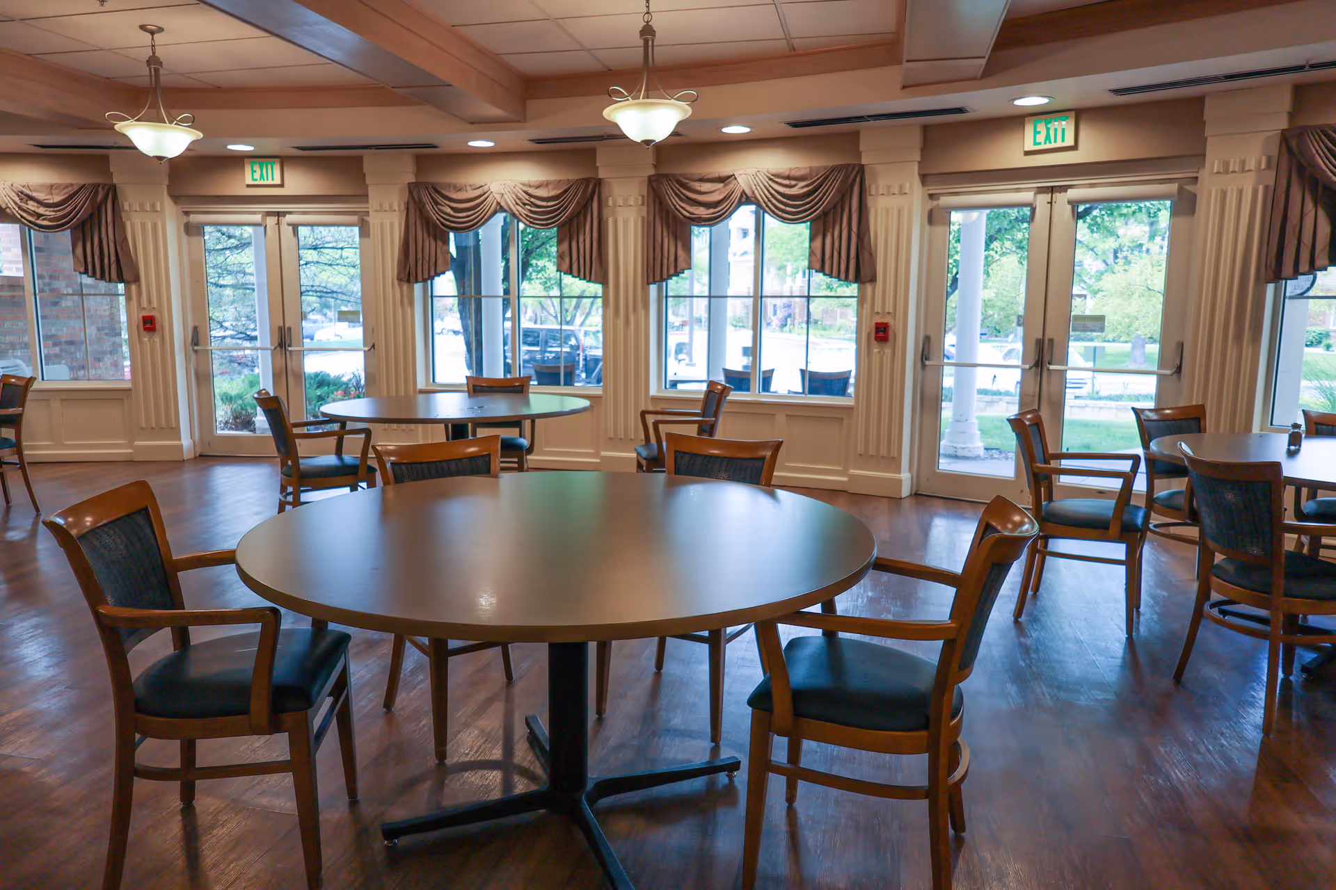 Communal dining room with round tables and wooden chairs, large windows and glass doors letting in daylight.