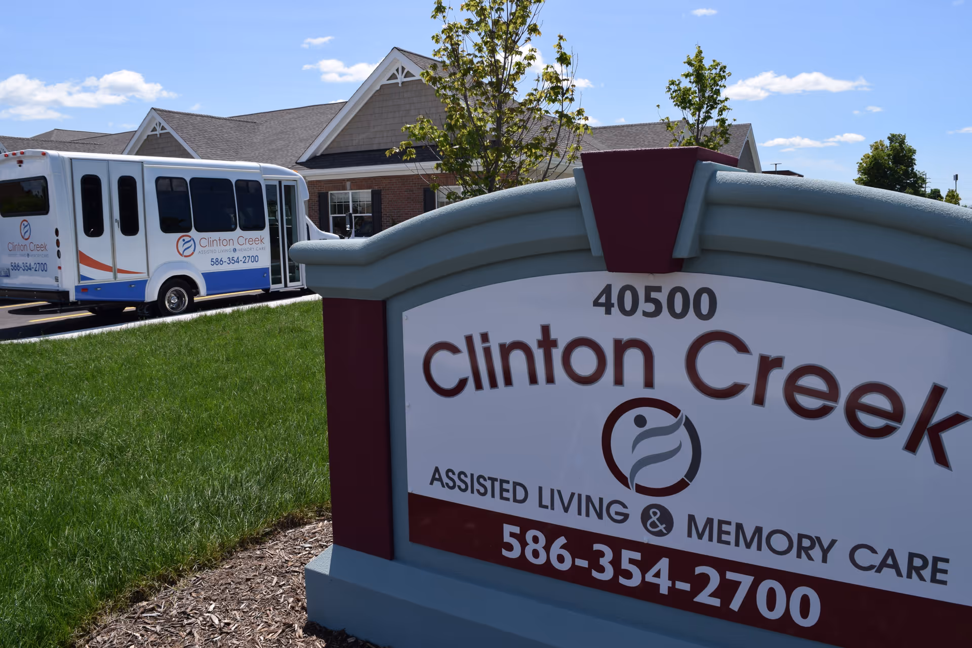 Outdoor view of Clinton Creek Assisted Living & Memory Care facility showing a large sign with the facility's name, address, and phone number in the foreground, and a white shuttle bus with the same branding parked near a brick building under a blue sky with some clouds.
