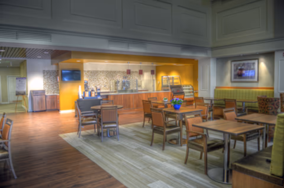 A spacious dining area in an assisted living facility featuring multiple wooden tables and chairs arranged on a carpeted section of the floor. The background includes a serving counter with a mosaic tile backsplash, beverage dispensers, and a small display case. The walls are painted in warm tones with framed artwork and soft lighting, creating a welcoming atmosphere.