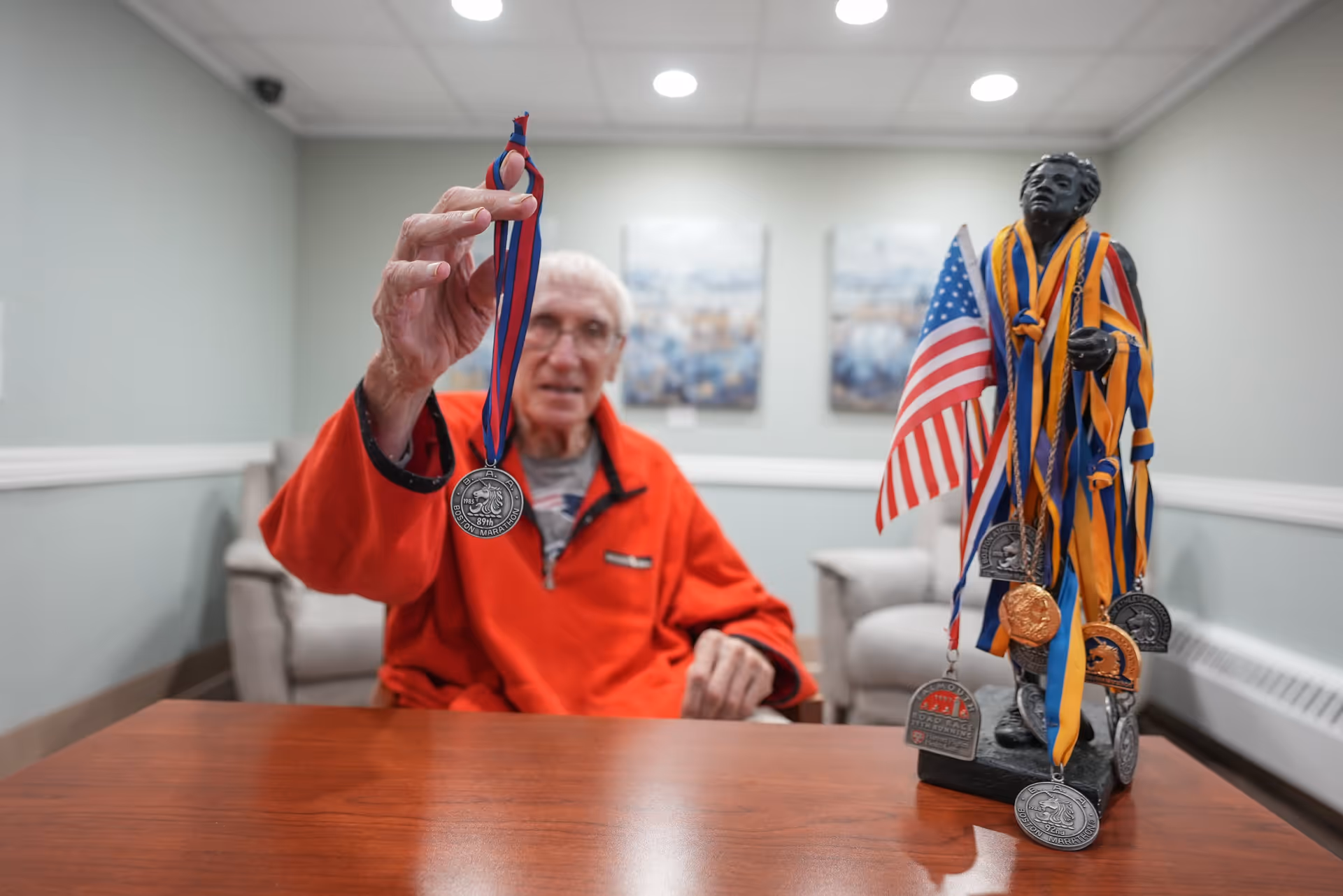 An elderly man wearing a red jacket is sitting at a wooden table in a room with light green walls and ceiling lights. He is holding up a medal with a blue and red ribbon. On the table in front of him is a statue draped with multiple medals and an American flag. Two framed pictures are visible on the wall behind him.