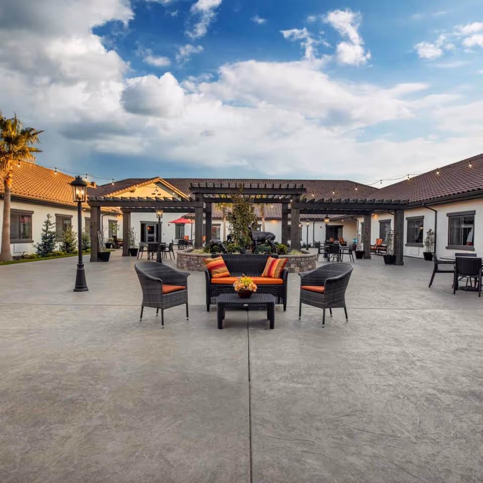Outdoor courtyard area of a senior living facility with a seating arrangement consisting of a black wicker loveseat and two chairs with orange cushions around a small table with a flower arrangement. The courtyard features a central stone planter with greenery and deer sculptures, surrounded by a wooden pergola. The building has a tiled roof and white walls, with additional tables and chairs scattered around the courtyard. The sky is partly cloudy with blue patches visible.
