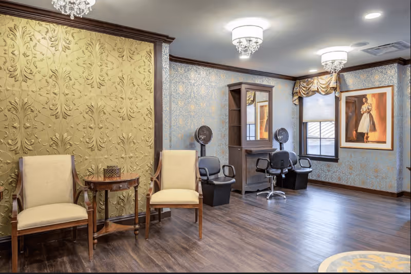 Interior of a salon area in a senior living facility with two beige chairs and a small wooden table against a green textured wall on the left. On the right side, there are two black salon chairs with hair dryers and a wooden styling station with a mirror. The walls have blue patterned wallpaper, and there is a framed painting of a woman in a dress near a window with a valance. The floor is dark wood, and two ceiling light fixtures with crystal accents are visible.