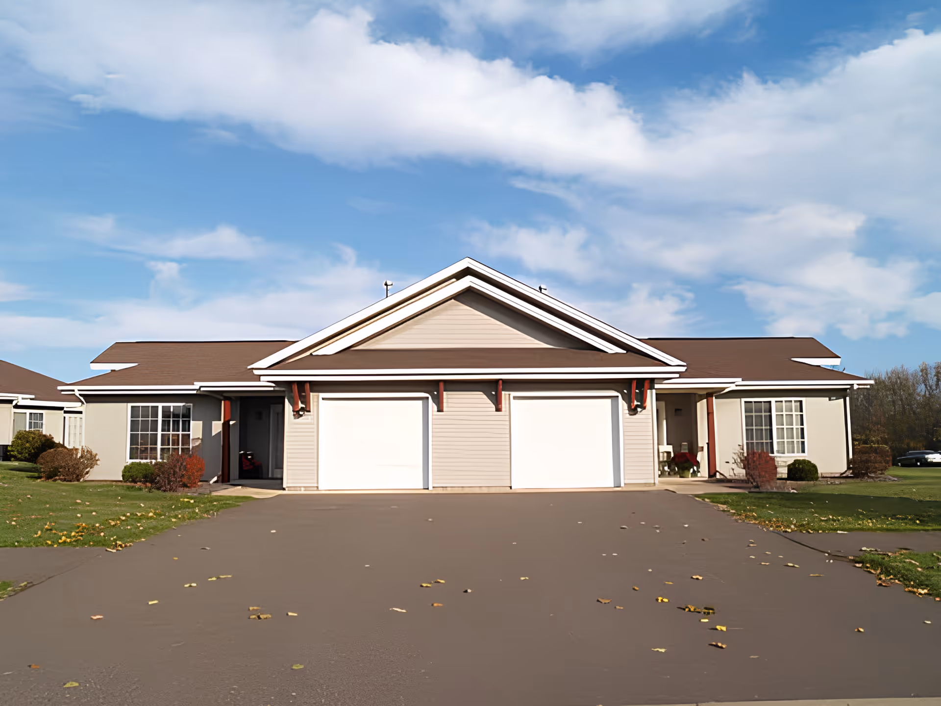 Front exterior view of a single-story building with two garage doors, beige siding, brown roof, and a driveway leading up to it. There are windows on either side of the garages and some bushes and grass around the building under a partly cloudy blue sky.