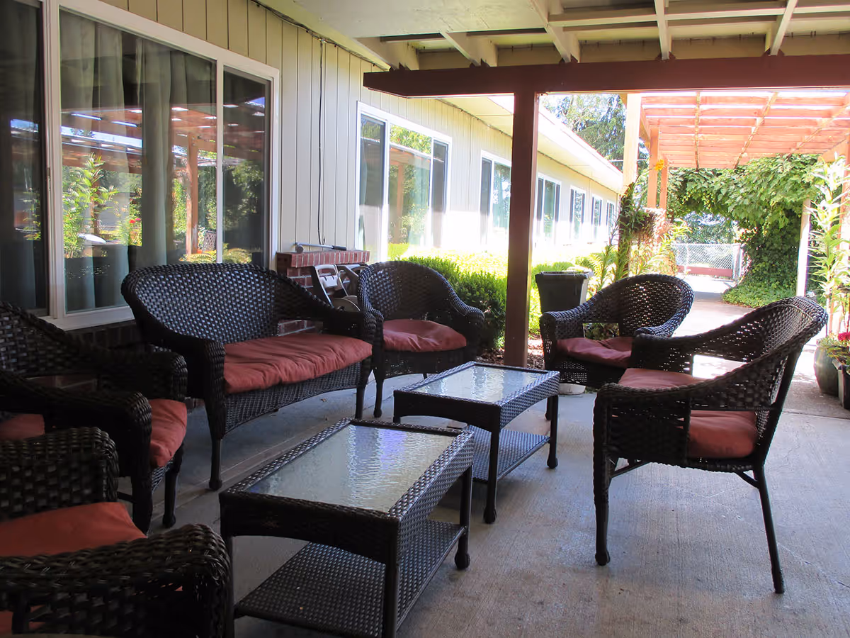 Outdoor covered patio area with wicker chairs and loveseats featuring red cushions, two glass-top coffee tables, and surrounding greenery including potted plants and vines on a trellis.