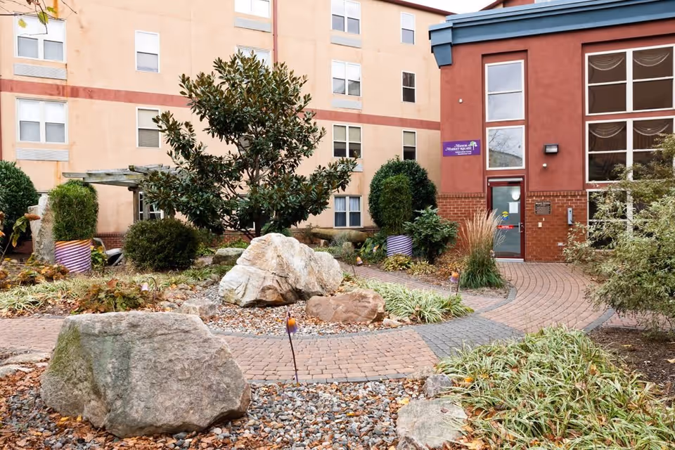 Outdoor courtyard area at The Manor at Market Square featuring a brick pathway winding through landscaped rocks, bushes, and trees, with a multi-story building in the background.