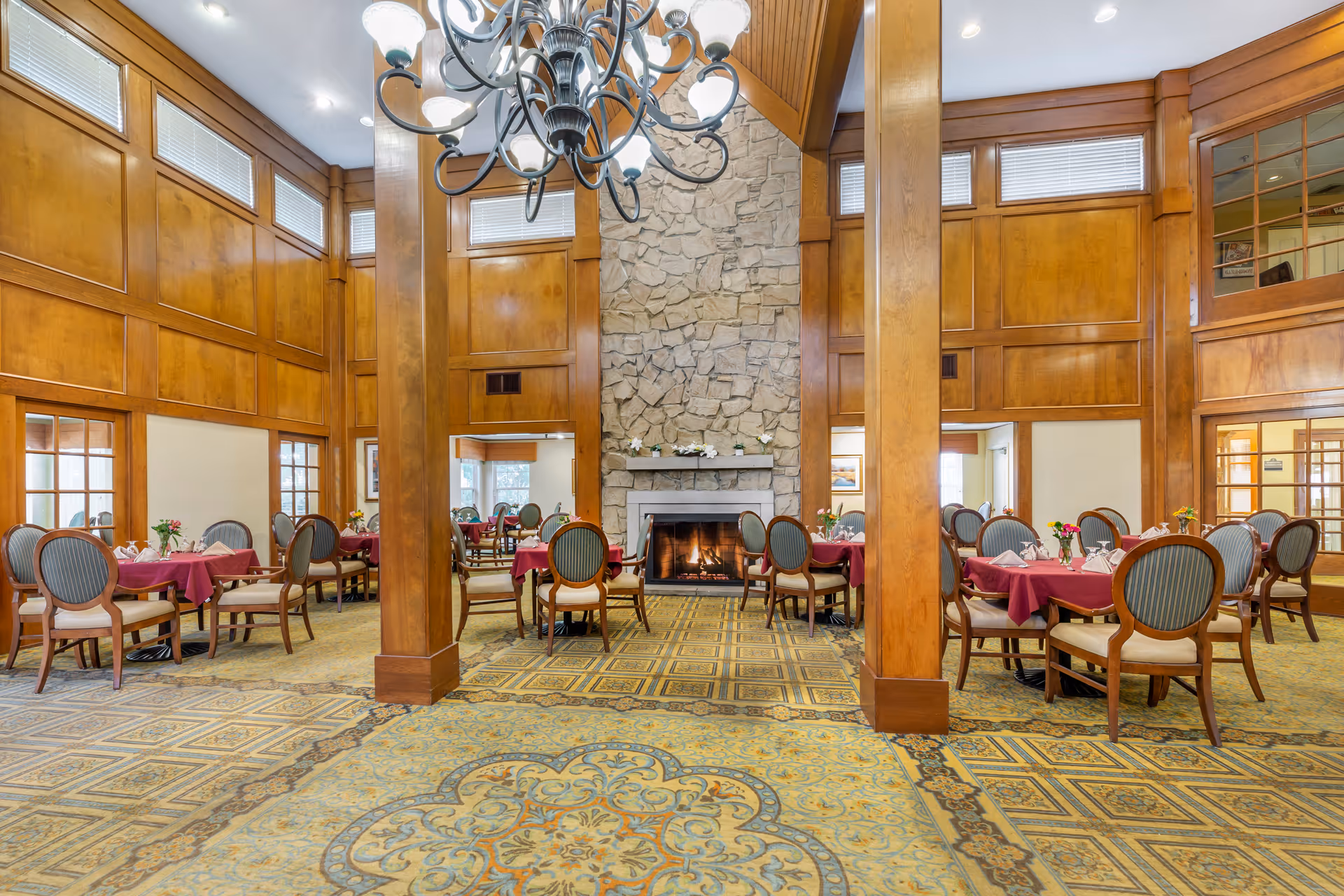A spacious dining room with multiple tables covered in burgundy tablecloths, each set with napkins and small flower arrangements. The room features wooden paneling on the walls, large wooden pillars, a patterned carpet, and a stone fireplace with a fire burning. A decorative chandelier hangs from the ceiling, and windows near the ceiling allow natural light to enter.