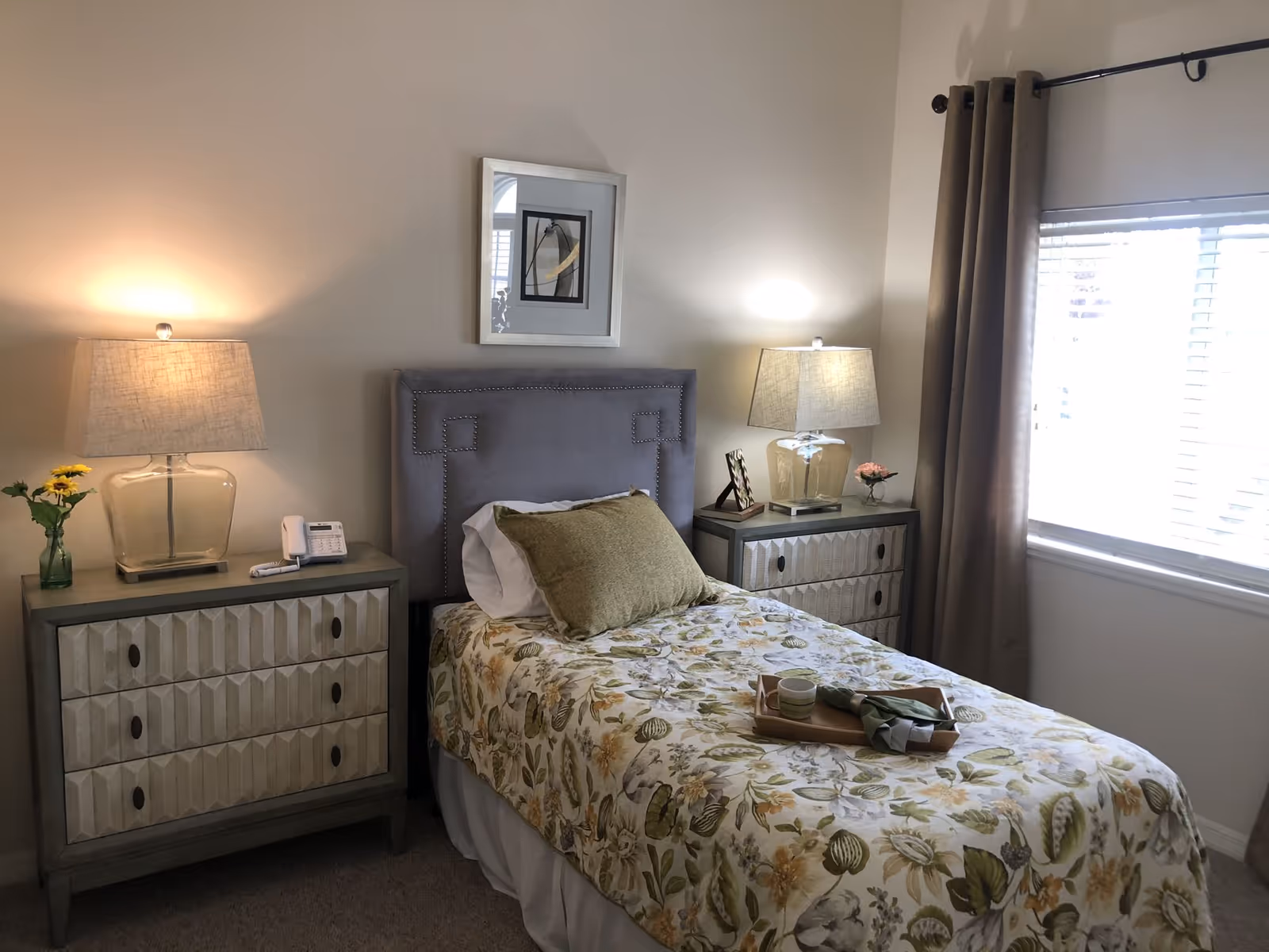A cozy bedroom with a single bed featuring a floral bedspread and a green pillow. There are two bedside tables on either side of the bed, each with a lamp and decorative items. A window with beige curtains allows natural light into the room, and a framed picture hangs above the bed.