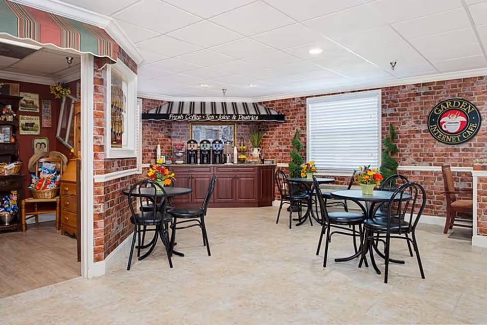 A cozy indoor seating area with several round tables and black metal chairs, each table decorated with a small flower arrangement. The walls are covered with red brick wallpaper, and there is a counter with coffee dispensers under a black and white striped awning that reads 'Fresh Coffee - Tea - Juice & Pastries'. A sign on the wall says 'Garden Internet Cafe'. The floor is tiled, and there is a window with white blinds.