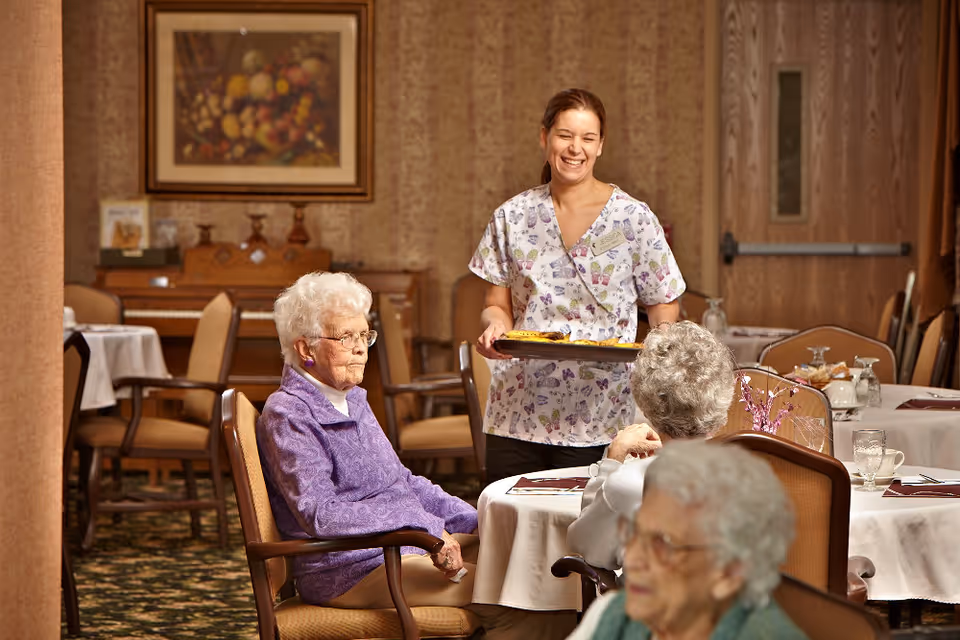 A smiling caregiver holding a tray with food stands next to a table where three elderly women are seated in a dining room with round tables covered in white tablecloths and wooden chairs. The room has warm lighting, a framed painting on the wall, and a piano in the background.