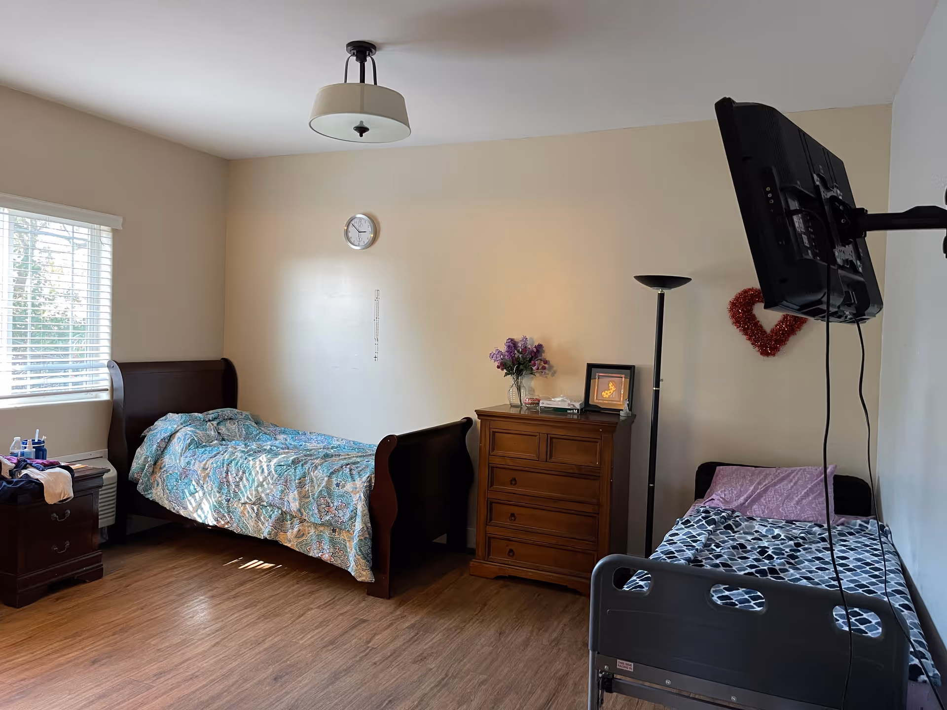 Shared bedroom with two beds, a wooden dresser, a wall-mounted TV and a window with blinds.