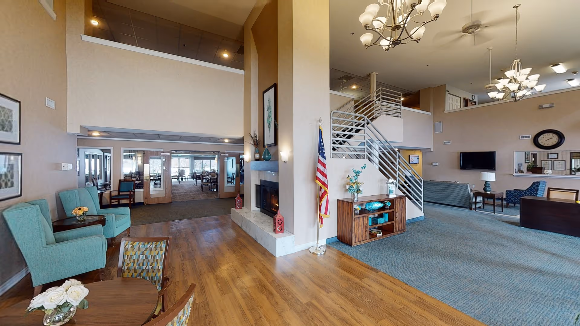Open two-story senior living lobby with seating areas, a fireplace, a staircase, and a dining area visible in the background.