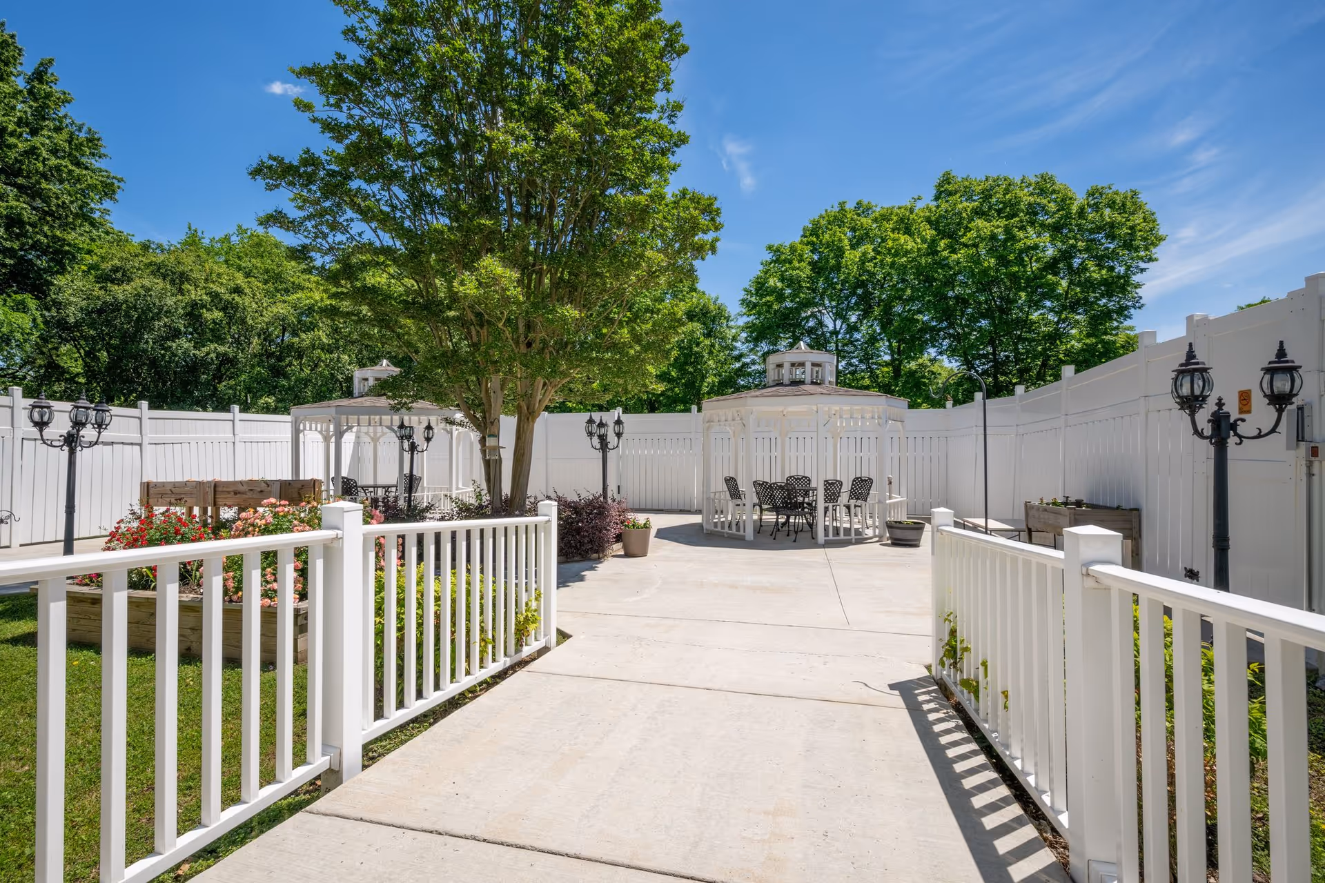 Outdoor courtyard area with white fencing, a concrete walkway, two white gazebos with black metal chairs and tables, surrounded by green trees and plants under a clear blue sky.
