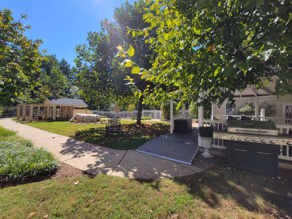Outdoor garden area with a paved walkway, green grass, trees, a large rock, a small shed, a metal chair, and a covered patio with a barbecue grill and potted plants under a clear blue sky.