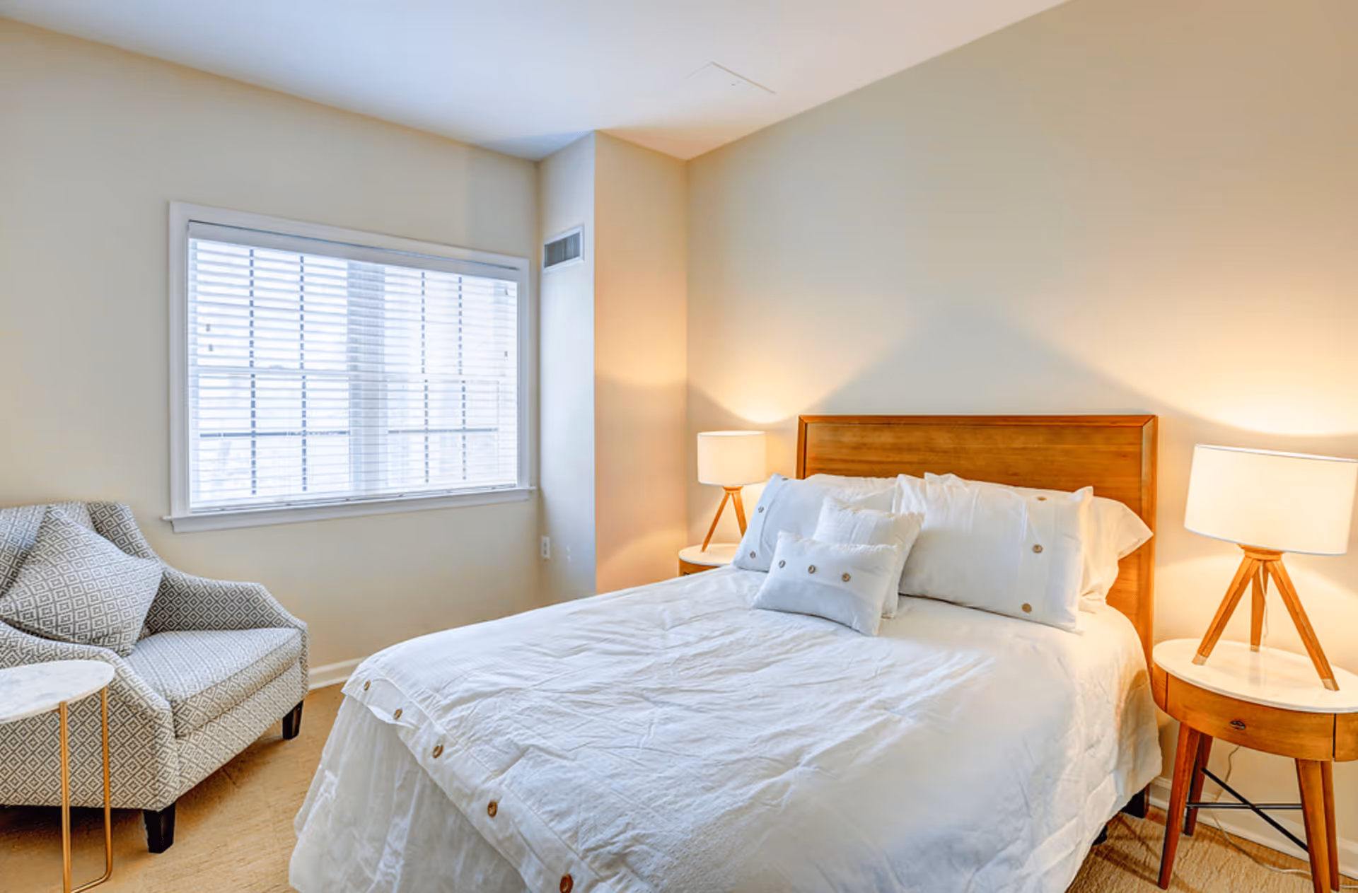 A cozy bedroom with a wooden bed frame, white bedding, and multiple pillows. There are two wooden nightstands with matching lamps on either side of the bed. A patterned armchair and a small round side table are positioned near a window with white blinds, allowing natural light to fill the room.