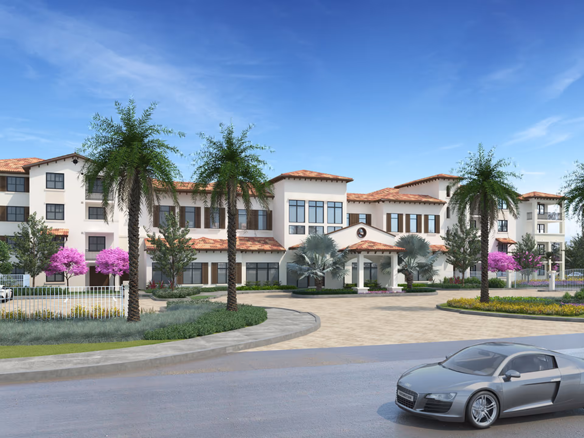 Front exterior of a Mediterranean-style senior living building with palm trees, landscaped circular drive and a silver car in the foreground.