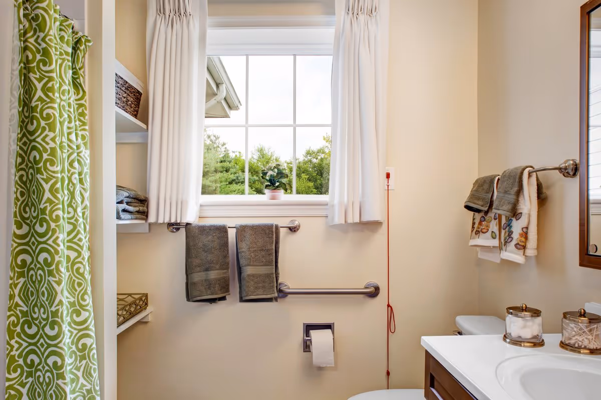 A bright bathroom with a window showing green trees outside. The bathroom features a green and white patterned shower curtain, white walls, and a white countertop with a sink. There are two gray towels hanging on a bar below the window, and additional towels hanging on a rack above the toilet. The countertop holds two glass containers with cotton balls and cotton swabs. A red emergency pull cord is visible on the wall near the toilet.