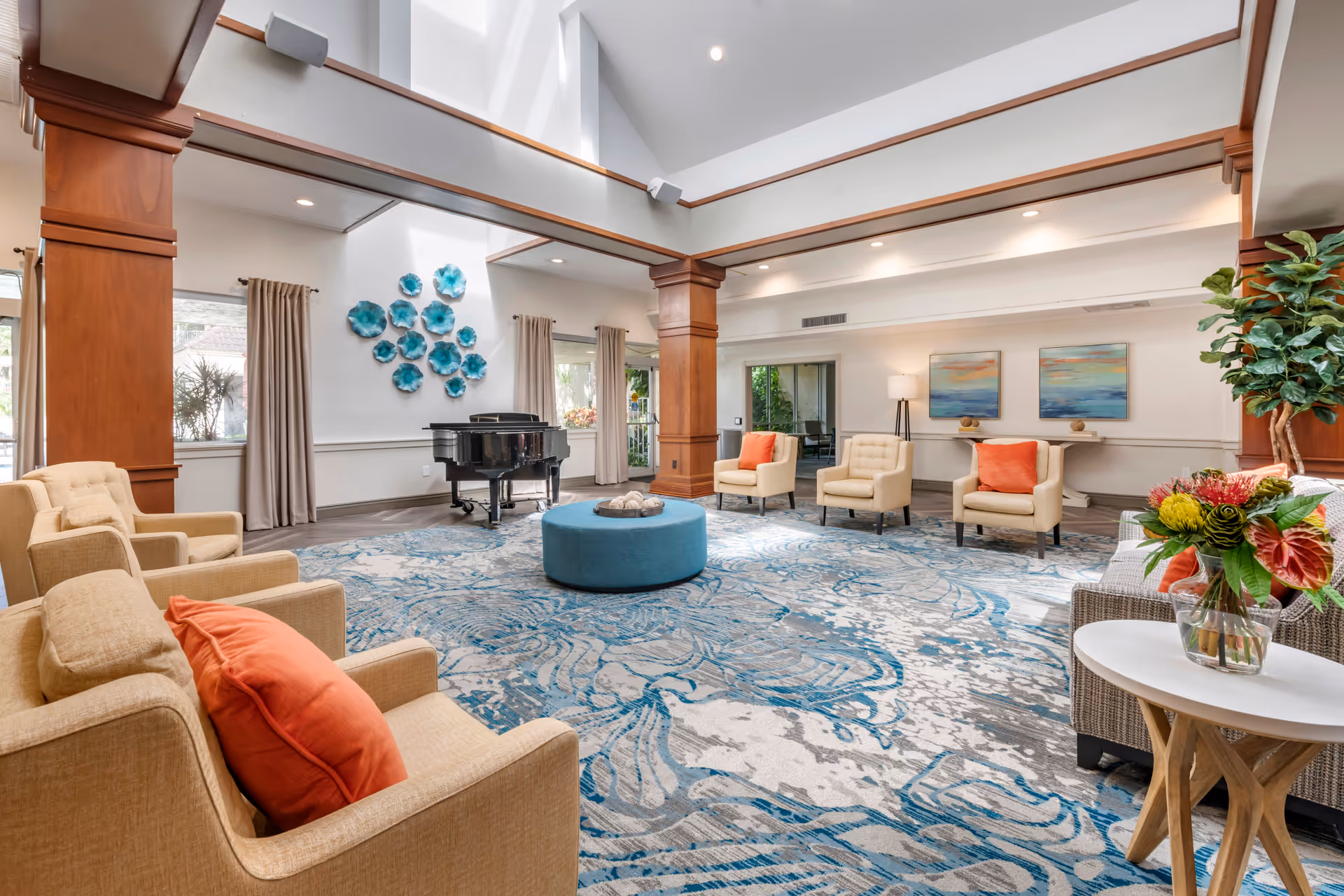 A spacious and well-lit living room area with beige armchairs featuring orange cushions arranged around a large blue and gray patterned rug. A round blue ottoman sits in the center, and a black grand piano is positioned against the far wall beneath a decorative wall installation of blue flowers. The room has high ceilings with wooden beams and large windows with beige curtains, allowing natural light to fill the space. A small table with a vase of colorful flowers is visible in the foreground.