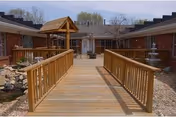Wooden bridge with railings leading to a courtyard area surrounded by single-story brick buildings. The courtyard features a small wooden gazebo structure and a multi-tiered water fountain. Trees and shrubs are visible in the background under a clear sky.