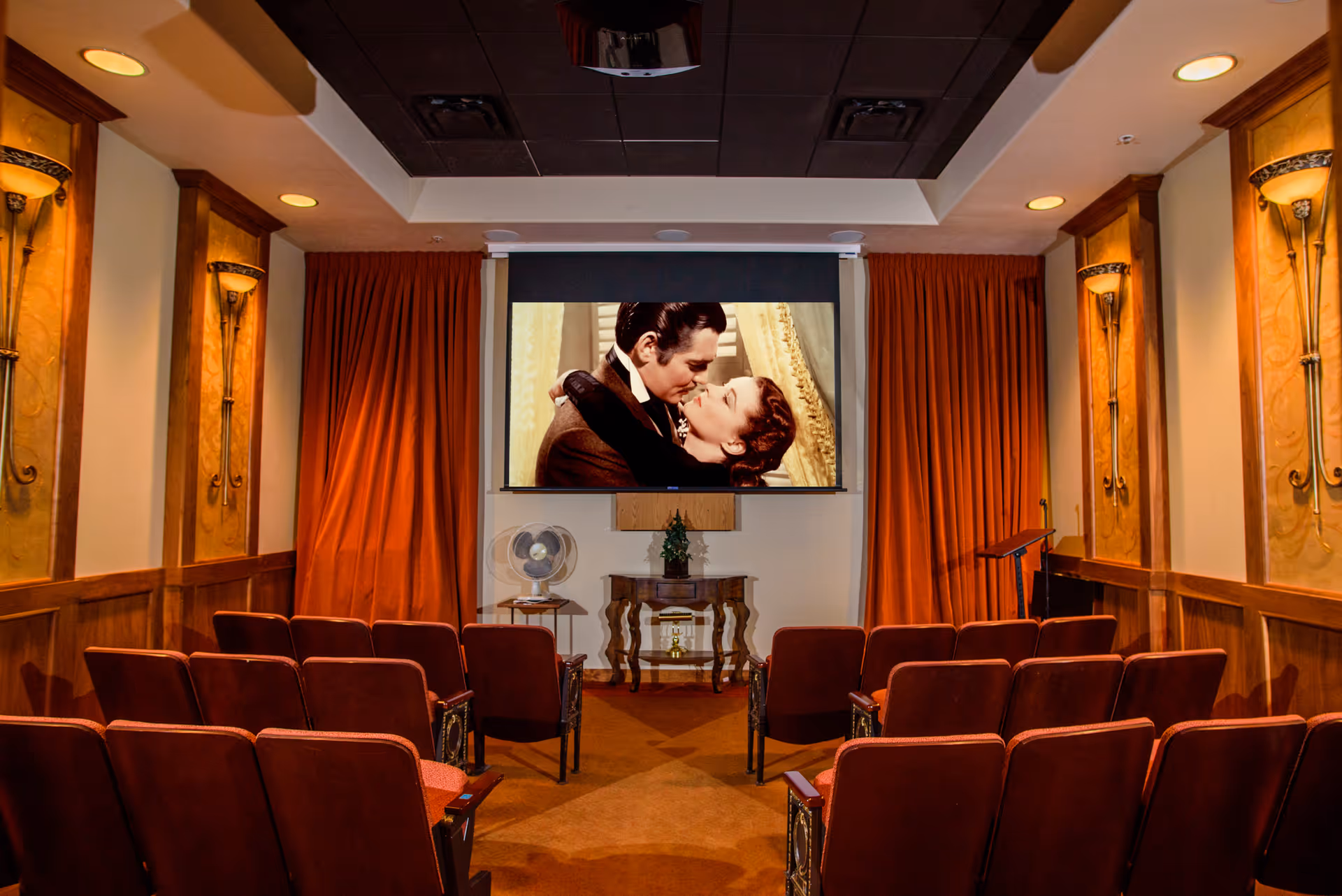 Small screening room with rows of red theater seats facing a projection screen showing a kissing couple, wood-paneled walls and red curtains.