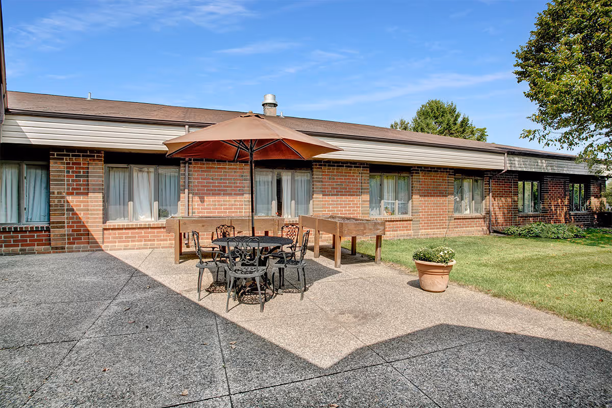 Outdoor patio area with a round metal table and four chairs under a large brown umbrella. The patio is adjacent to a single-story brick building with several windows. There are two raised garden beds and a potted plant on the patio. The area is surrounded by green grass and trees under a clear blue sky.